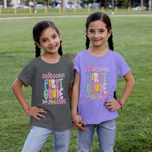 Two young girls wearing 'First Grade' t-shirts standing on a grassy field.