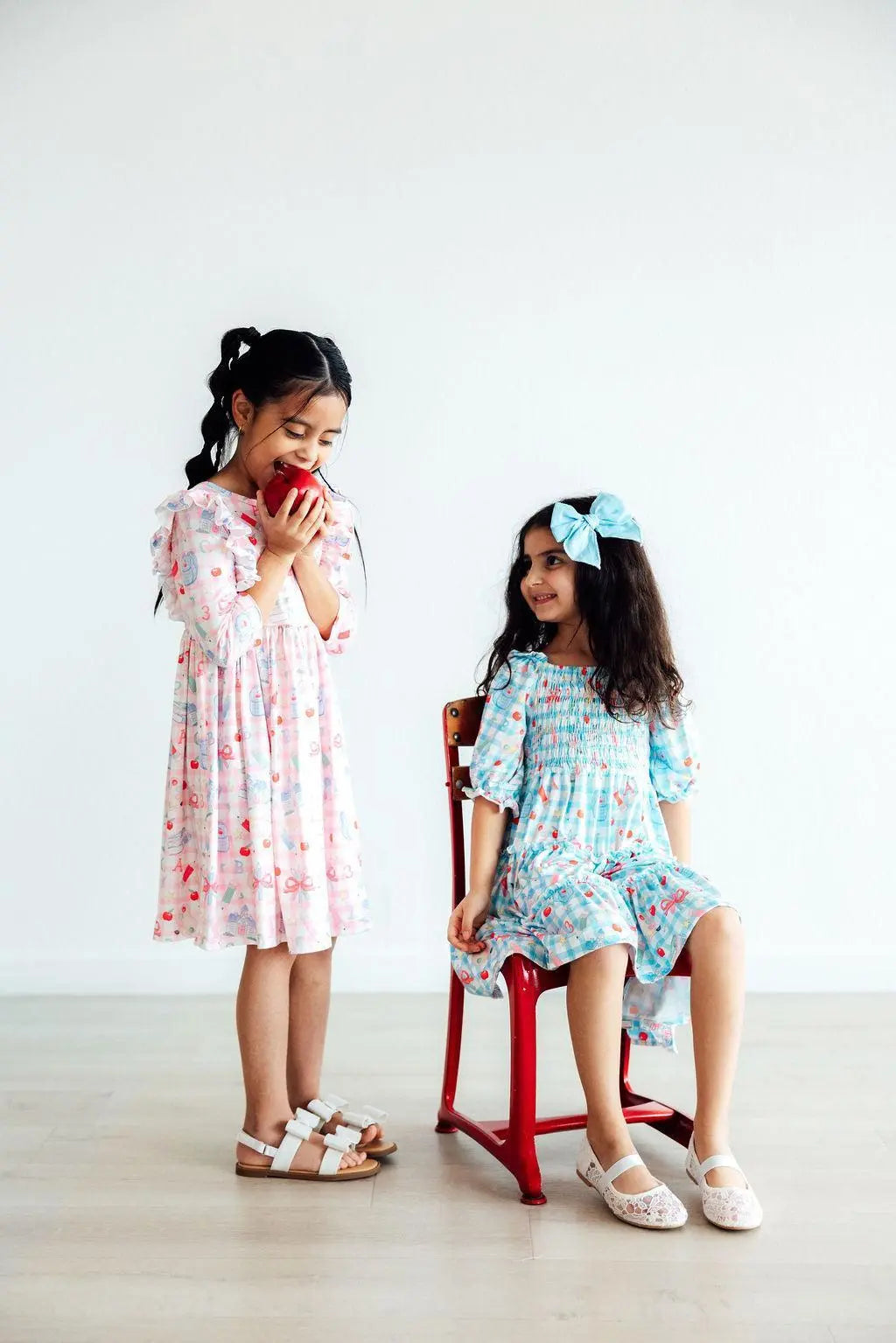 Two young girls in dresses standing and sitting on a stool against a white background