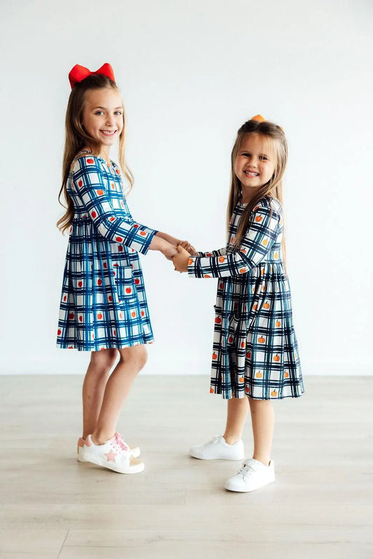 Two young girls wearing matching checkered dresses holding hands on a light wooden floor.