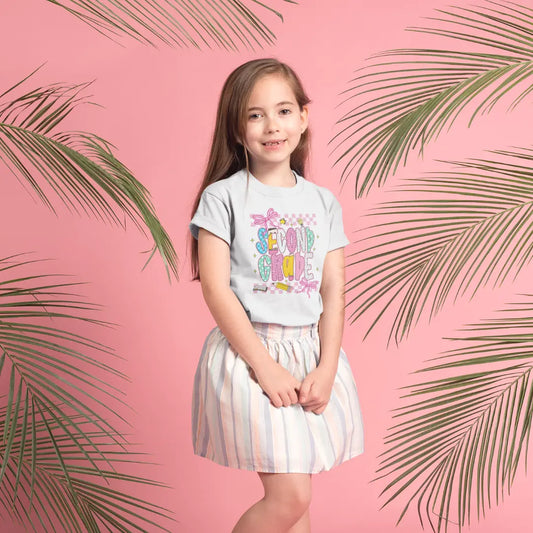 Young girl wearing a colorful second grade t-shirt with text and striped skirt on a pink background with palm leaves.