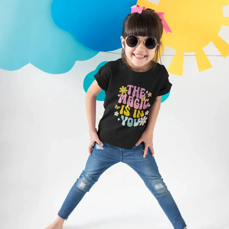 Child wearing a black t-shirt with colorful text, standing against a bright background with blue cloud and yellow sun shapes.