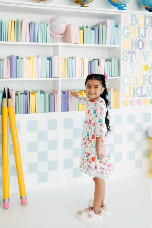 Child in a colorful dress standing in front of a bookshelf with pastel books and alphabet letters.