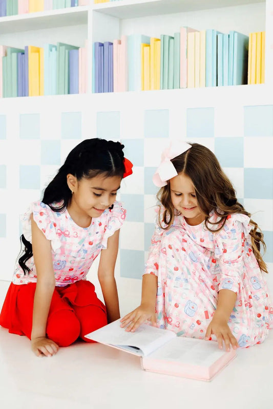 Two young girls in matching dresses reading a book in front of a colorful bookshelf.