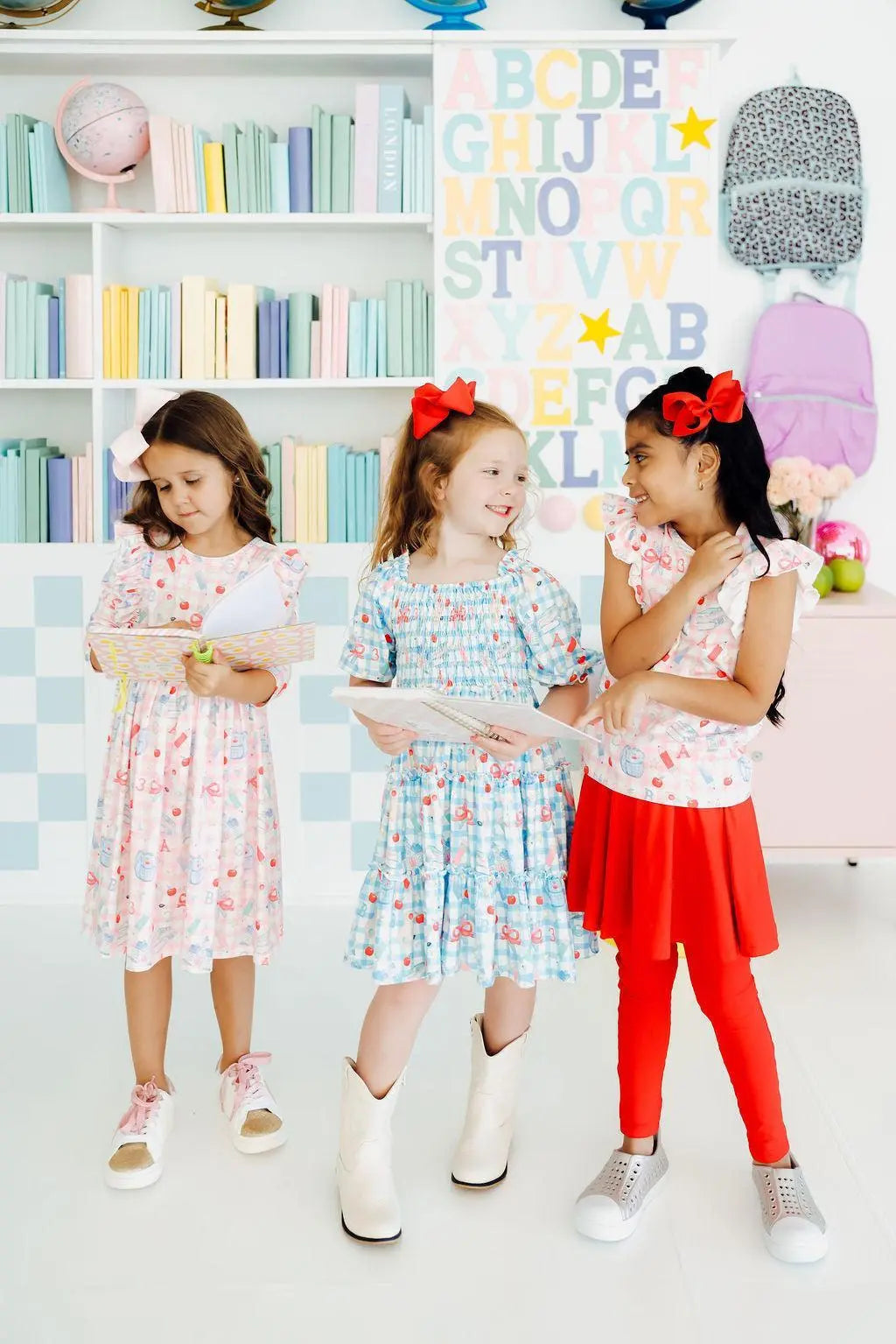 Three young girls in colorful dresses standing in a room with a bookshelf and alphabet poster.