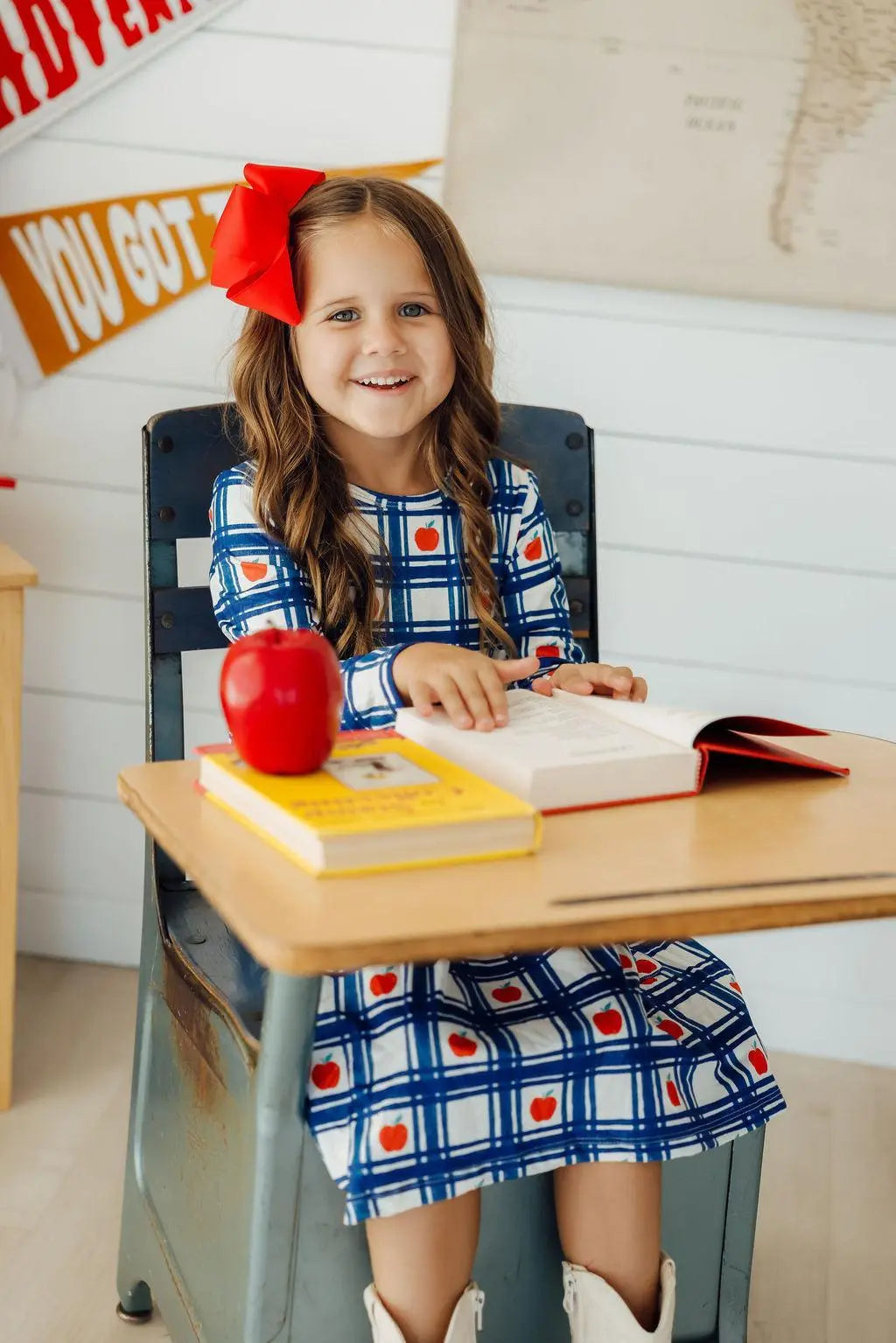Young girl sitting at a desk with books and an apple, wearing a red bow in her hair.