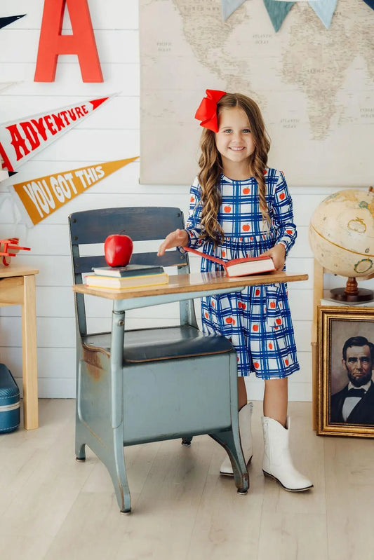 Young girl in a plaid dress with a red bow standing next to a vintage school desk in a classroom setting.
