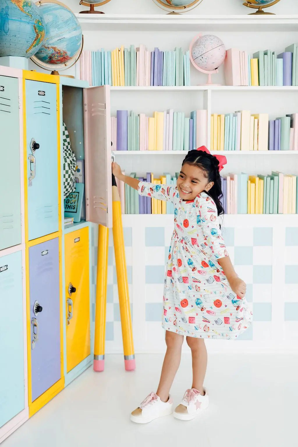 Young girl in a colorful dress standing in a room with pastel bookshelves and lockers.