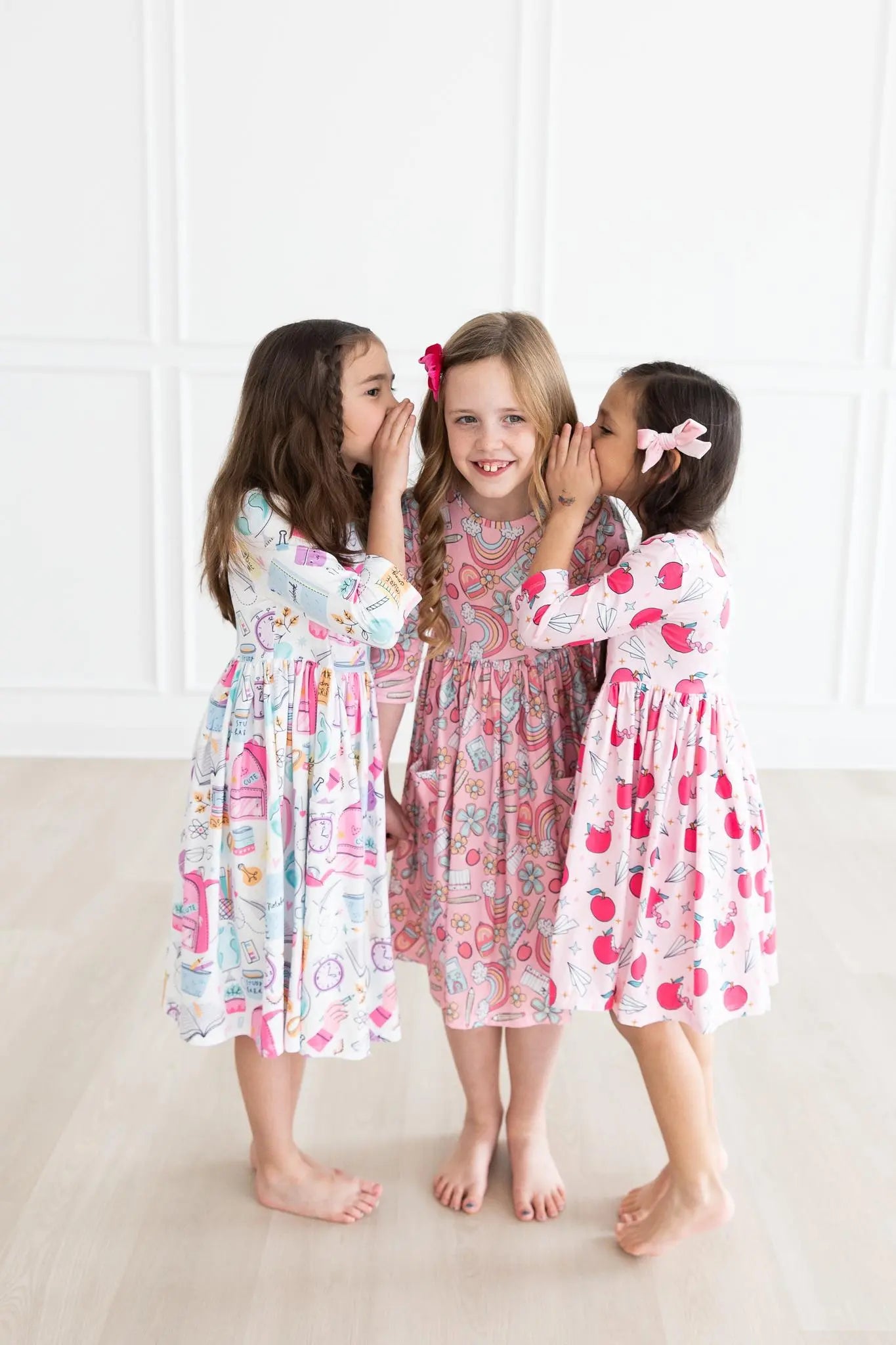 Three young girls in patterned dresses standing close together on a light-colored floor.
