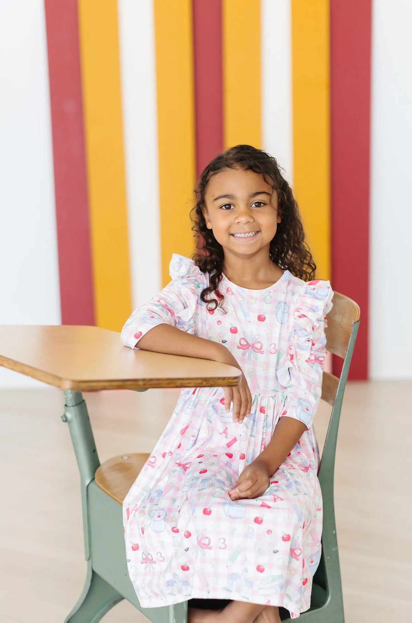 Young girl in a white dress with red patterns sitting at a table against a colorful striped wall.