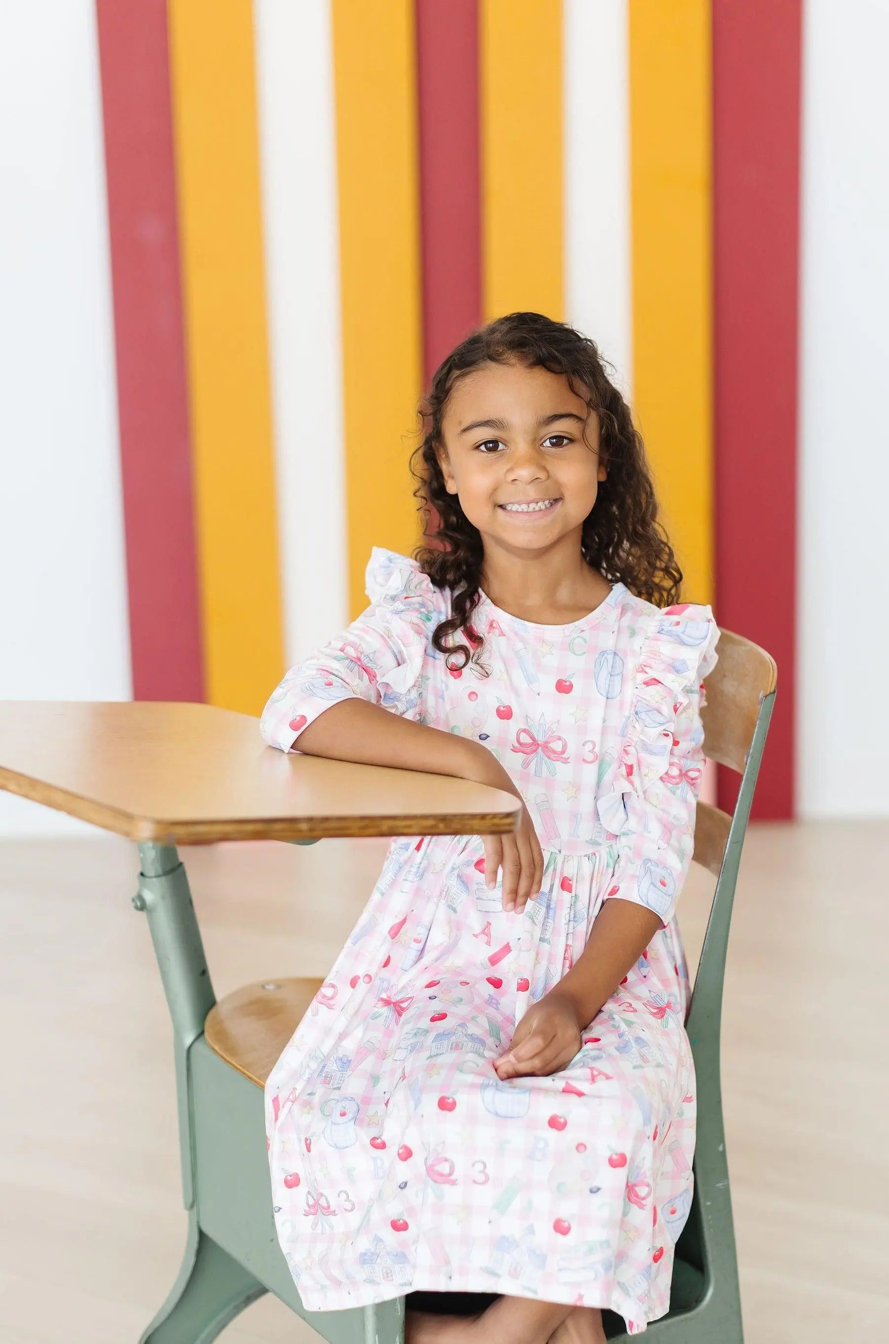 Young girl in a white dress with red patterns sitting at a table against a colorful striped wall.