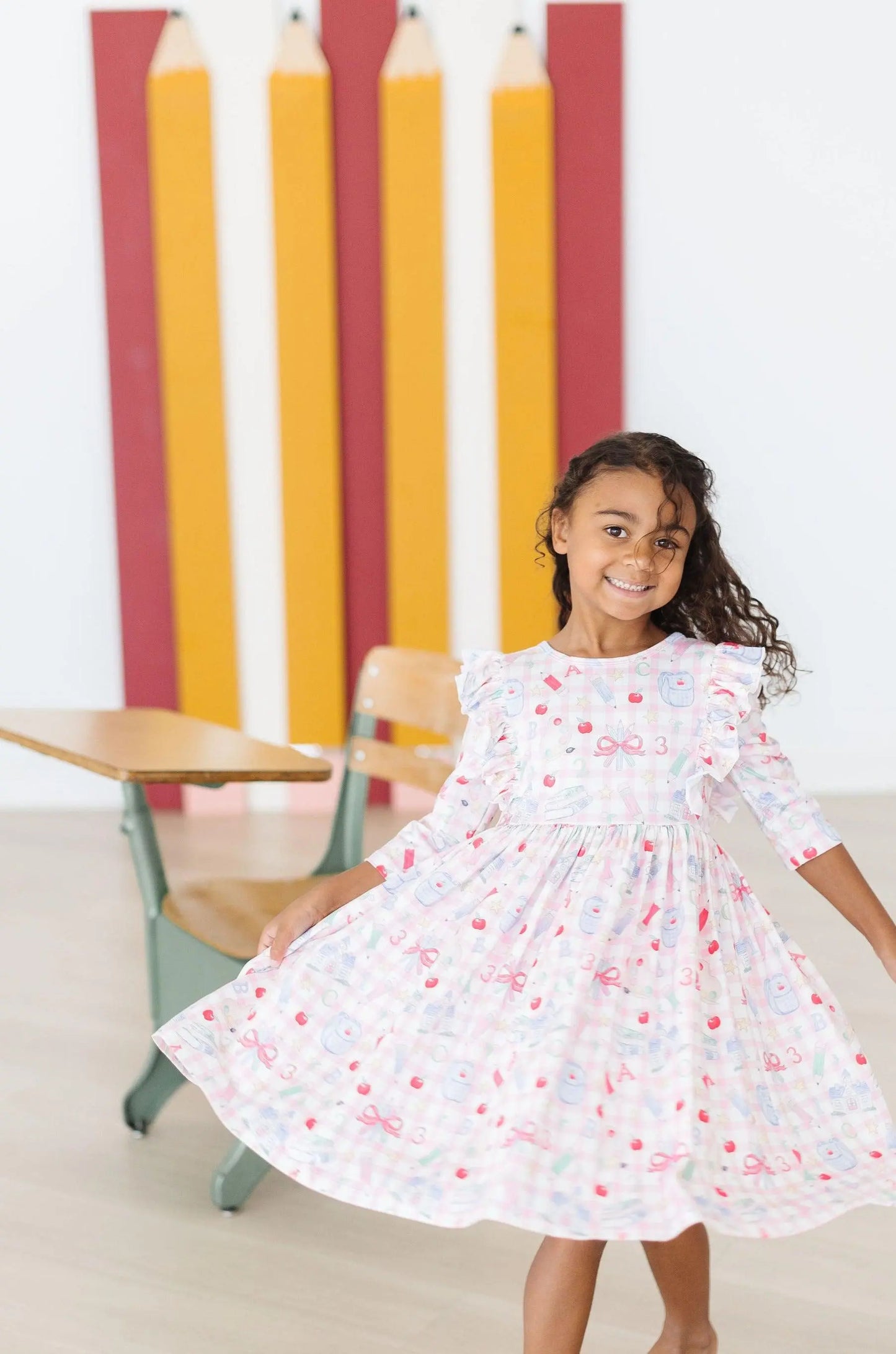Young girl in a floral dress standing in front of colorful pencils and a chair.