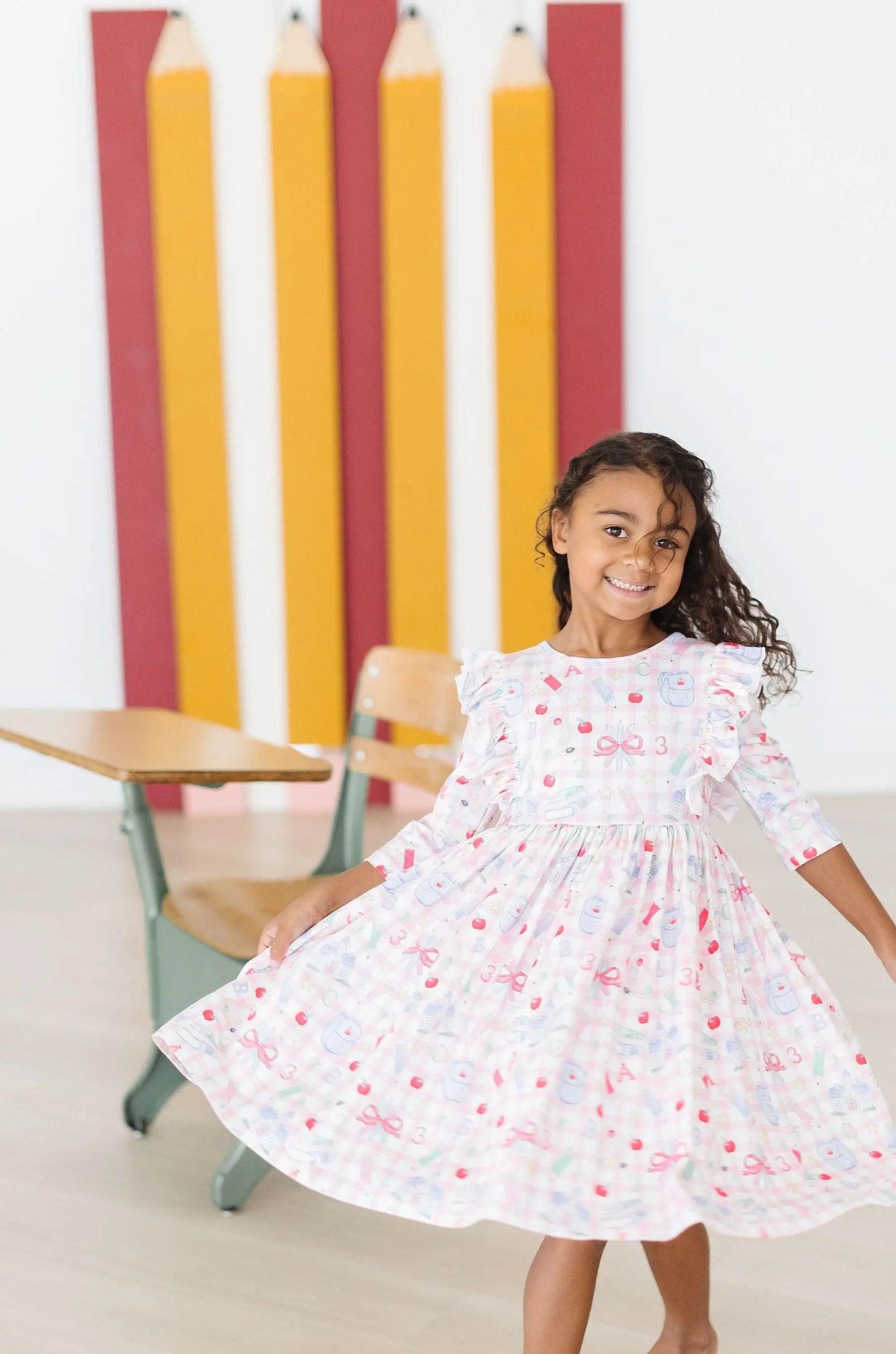 Young girl in a floral dress standing in front of colorful pencils and a chair.