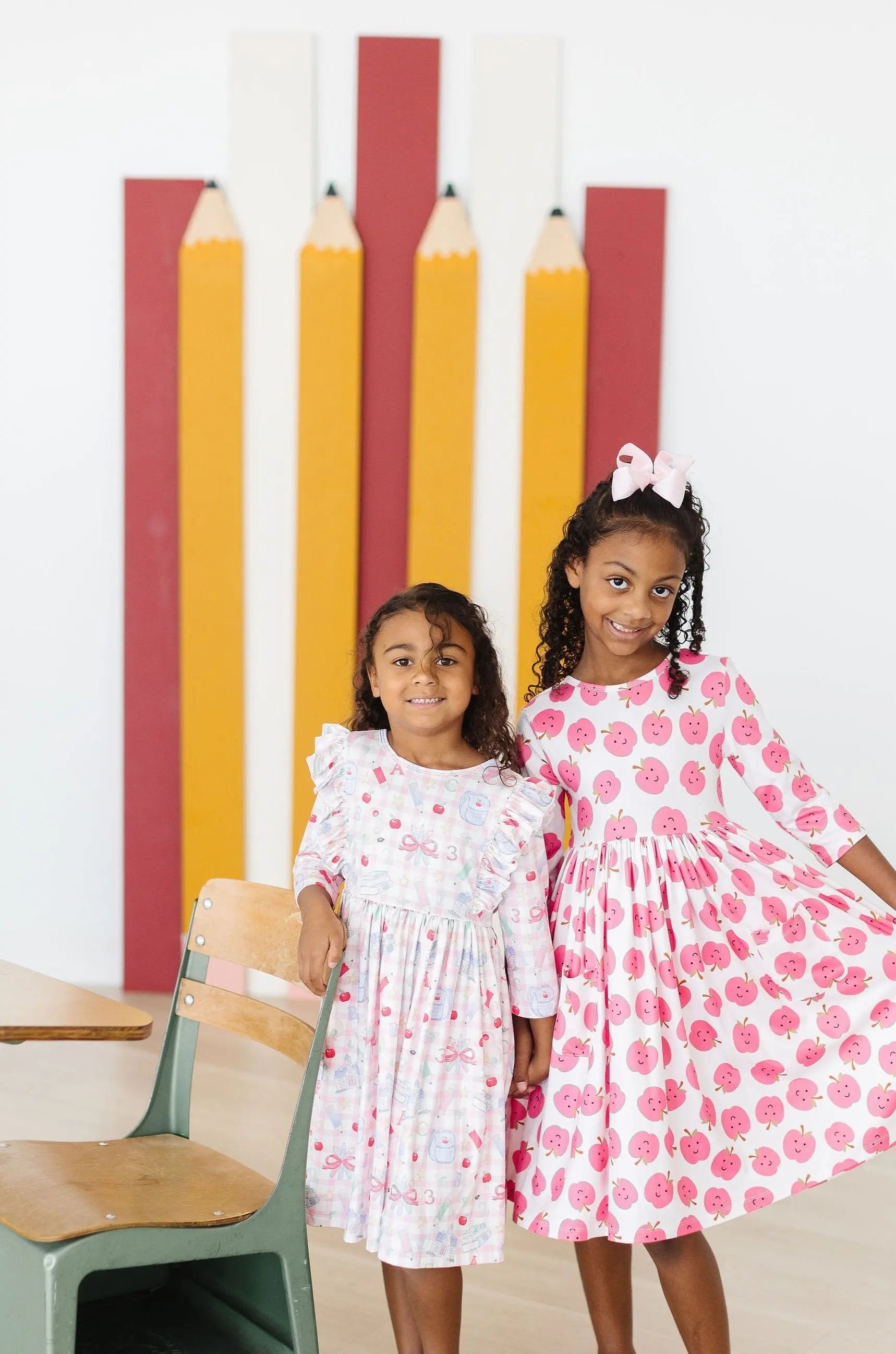 Two young girls in patterned dresses standing in front of a colorful pencil background