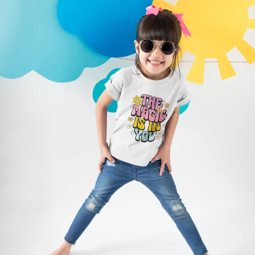 Child wearing a white t-shirt with colorful text and blue jeans, standing against a colorful cloud and sun background.