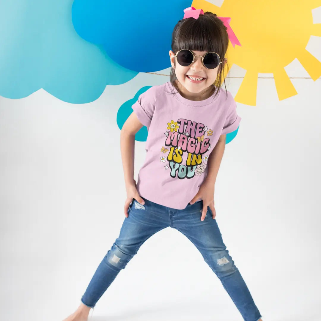 Child wearing a pink t-shirt with 'The Magic is in You' text, blue jeans, and sunglasses against a colorful cloud and sun background.