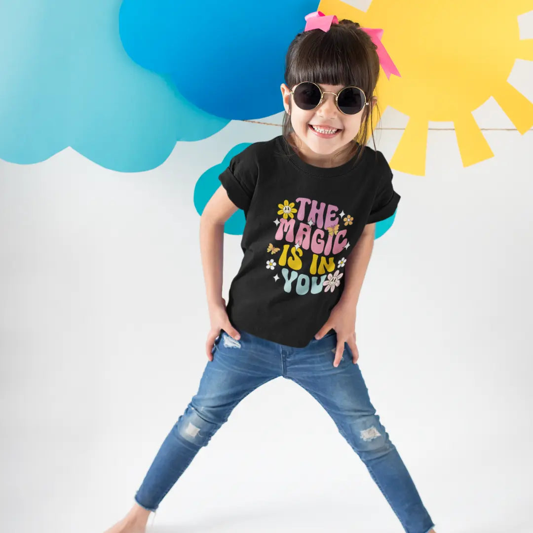 Child wearing a black t-shirt with colorful text, standing against a bright background with blue cloud and yellow sun shapes.