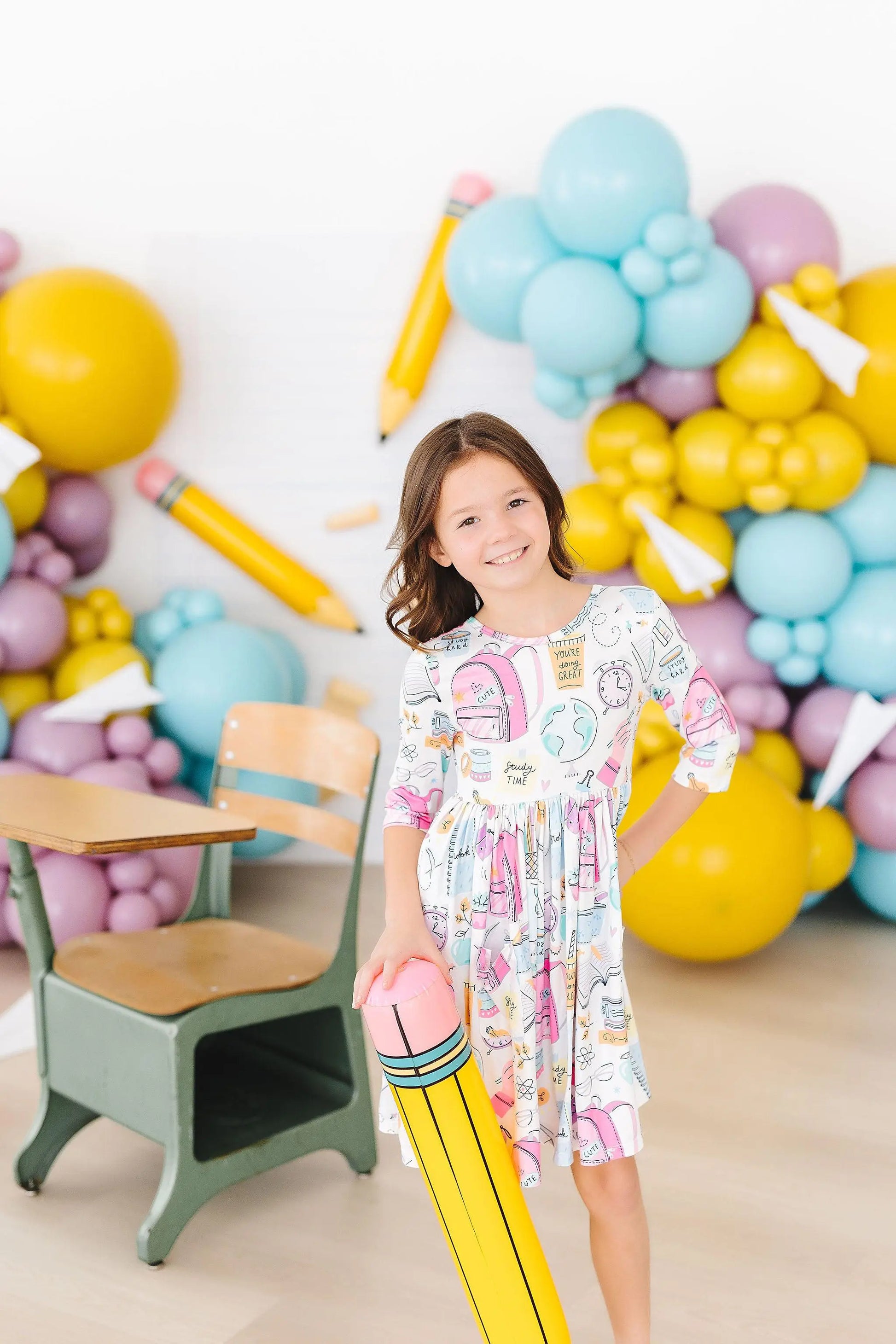 Young girl in a colorful dress standing in front of a wall decorated with balloons shaped like pencils and numbers.