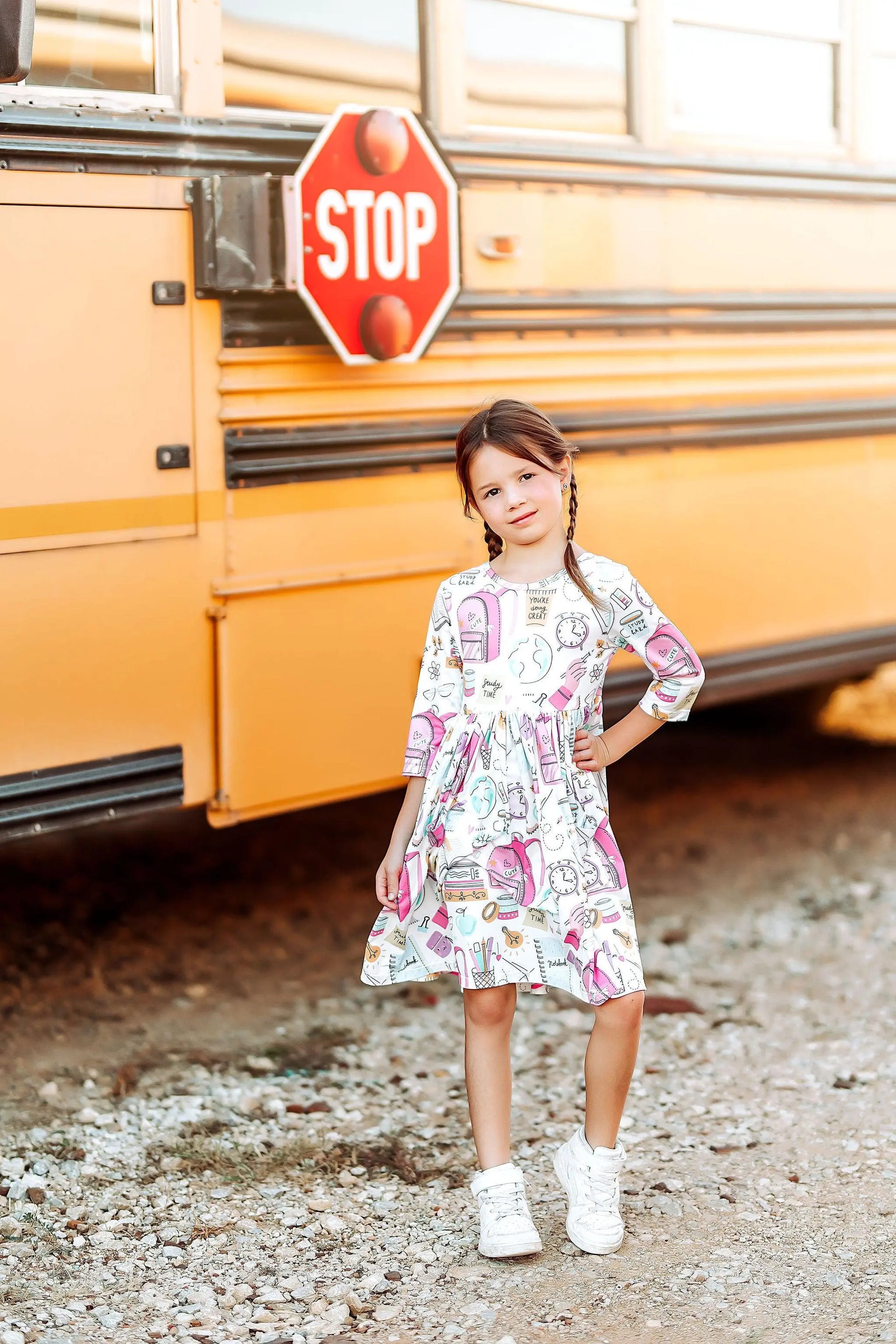 Young girl in a patterned dress standing in front of a yellow school bus with a stop sign.