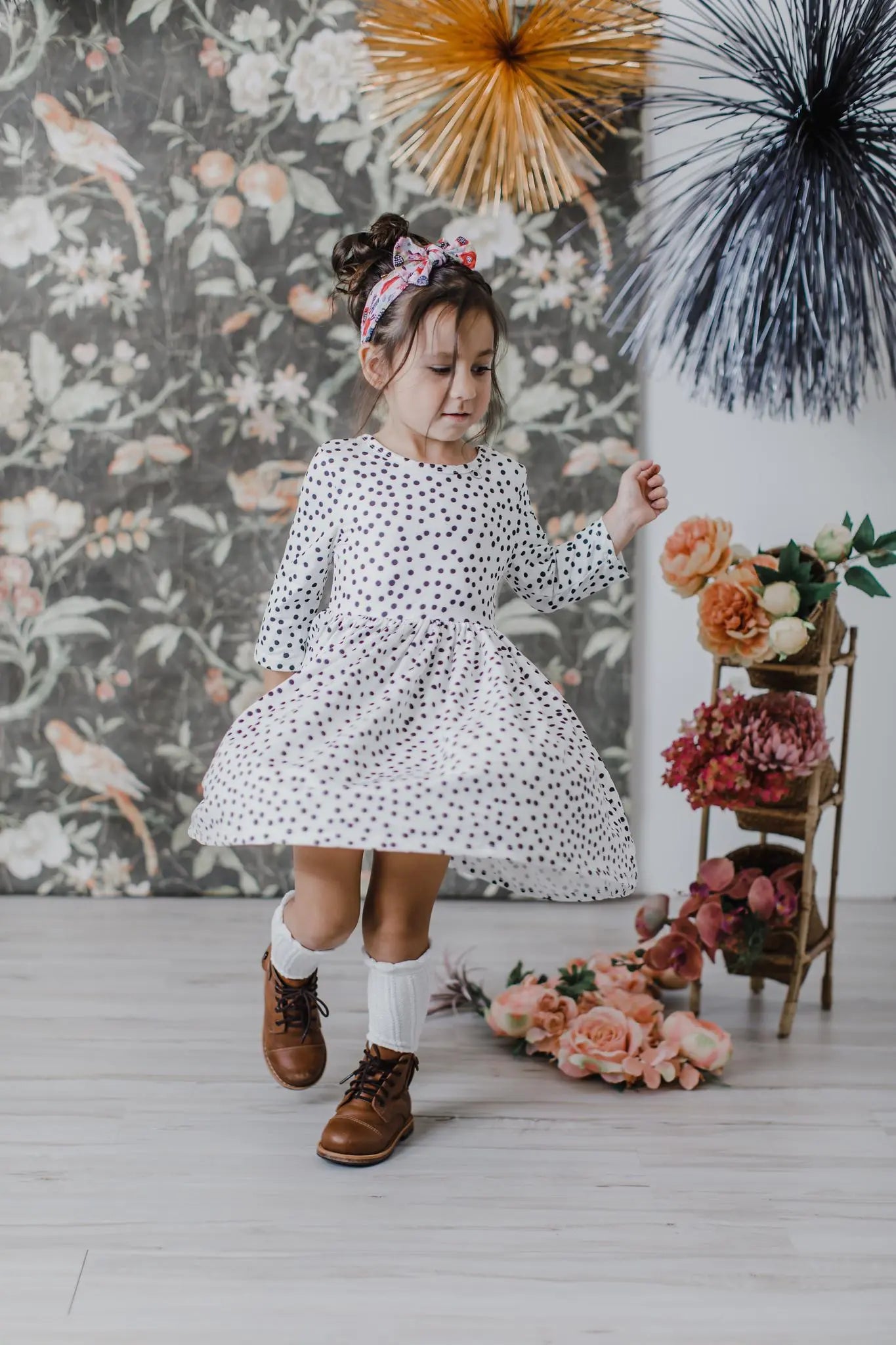 Young girl in a polka dot dress standing in a decorated room with floral decorations and a floral-patterned wall.