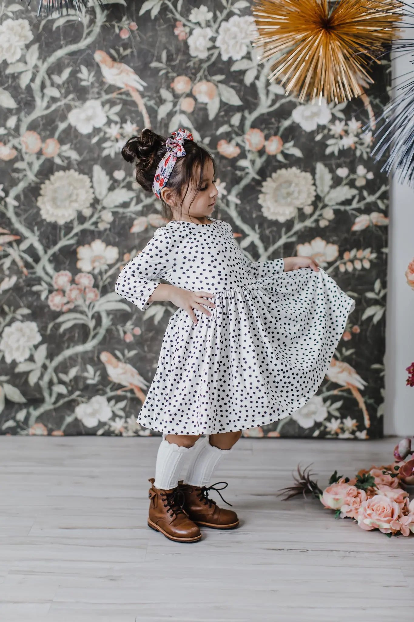 Child in a polka dot dress standing in front of a floral-patterned wall.