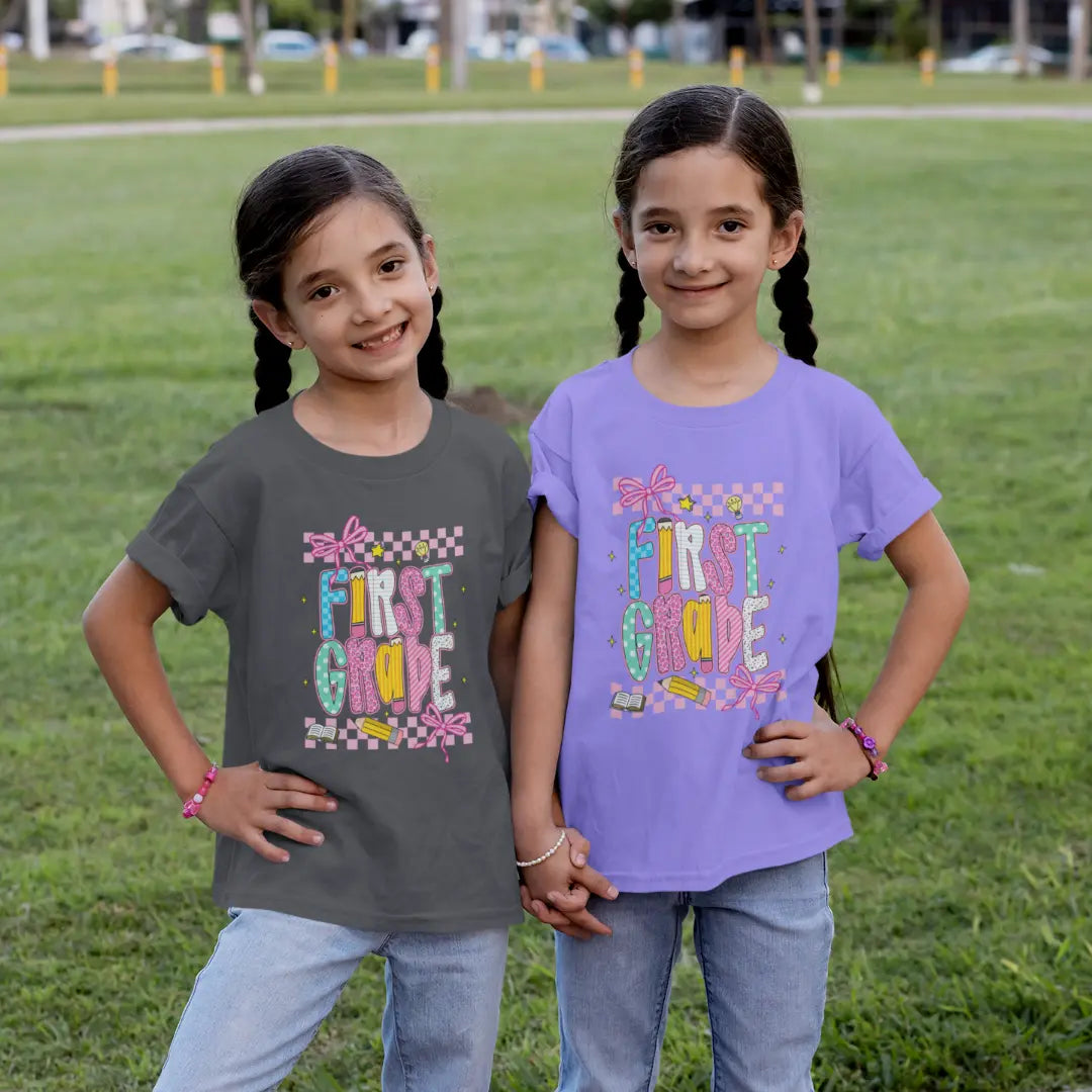 Two young girls wearing 'First Grade' t-shirts standing on a grassy field.