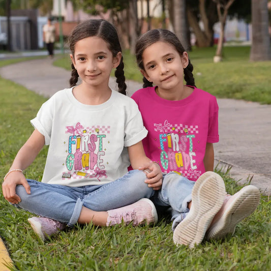 Two young girls sitting on grass wearing 'First Grade' t-shirts.