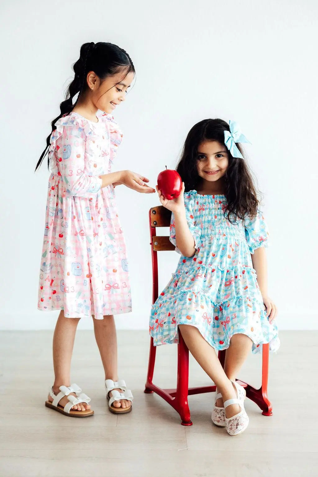 Two young girls in floral dresses interacting with an apple on a white background