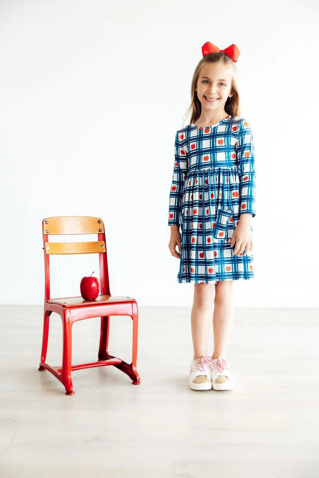 Young girl in a checkered dress standing next to a red chair with an apple on it.