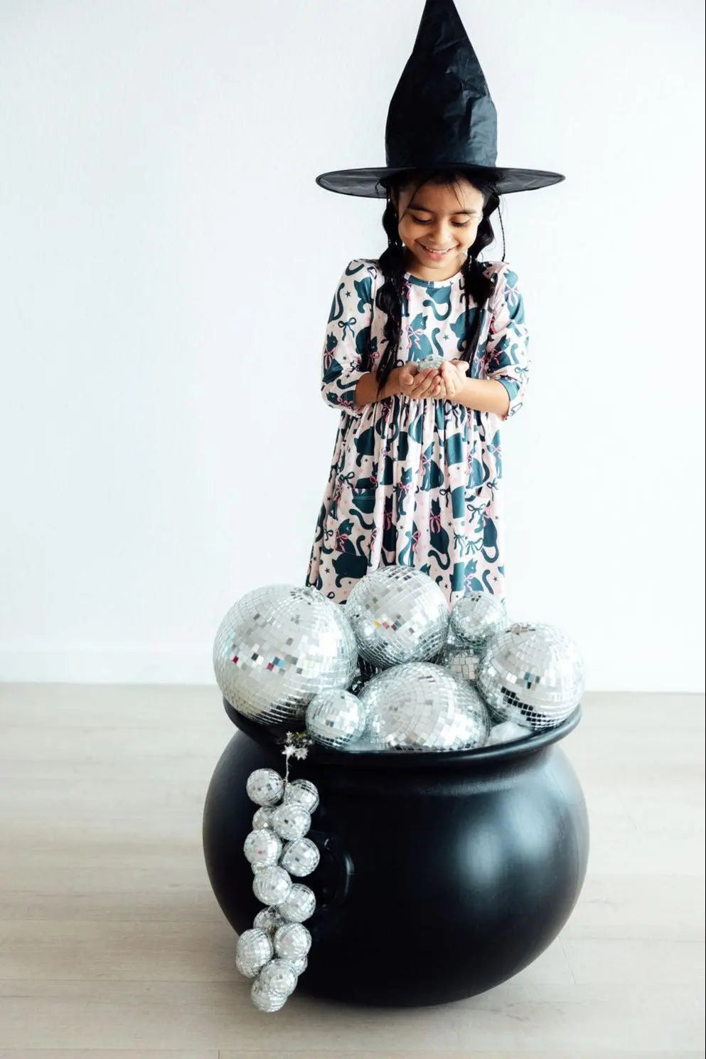 Child in a witch costume standing behind a black cauldron with silver decorations on a light background
