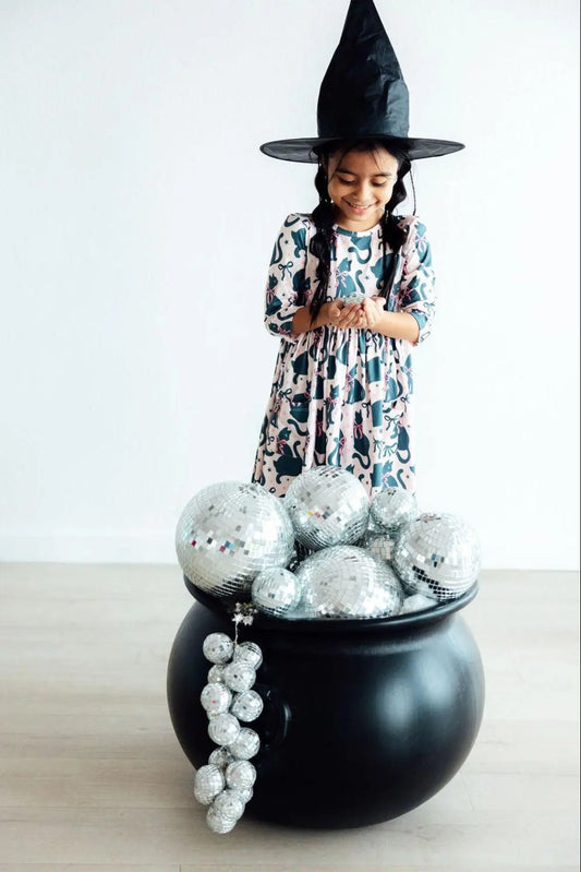 Child in a witch costume standing behind a black cauldron with silver decorations on a light background