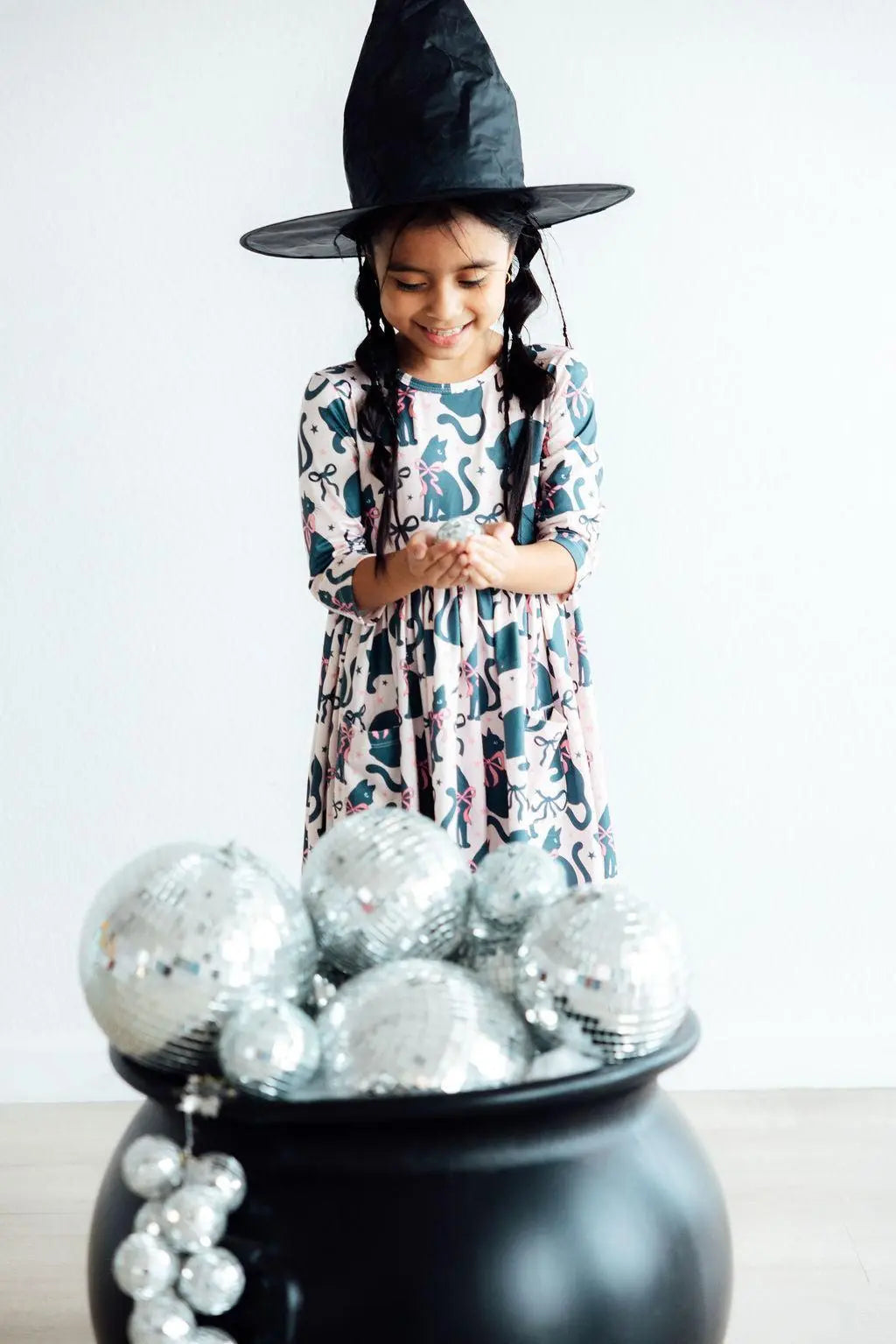 Child wearing a black witch hat and a patterned dress with cats and bows on a white background