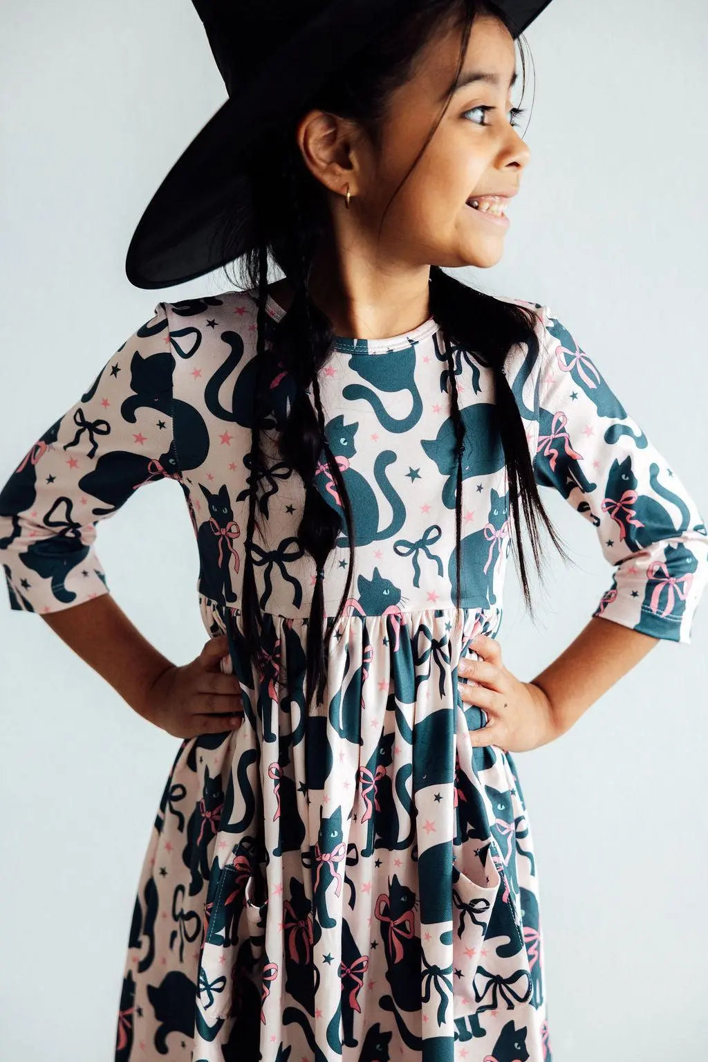 Child wearing a black witch hat and a patterned dress with cats and bows on a white background