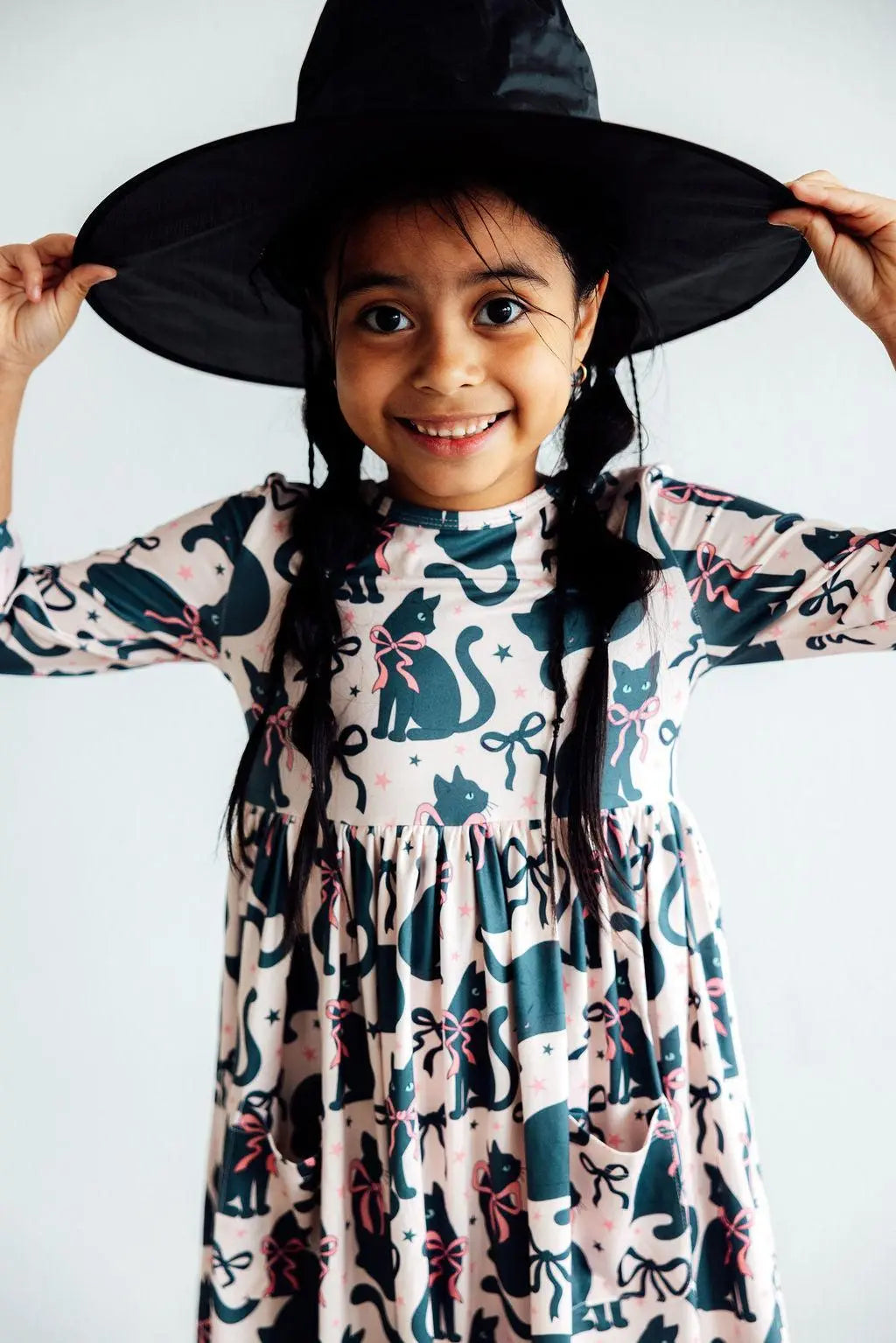 Child wearing a black witch hat and a patterned dress with cats and bows on a white background