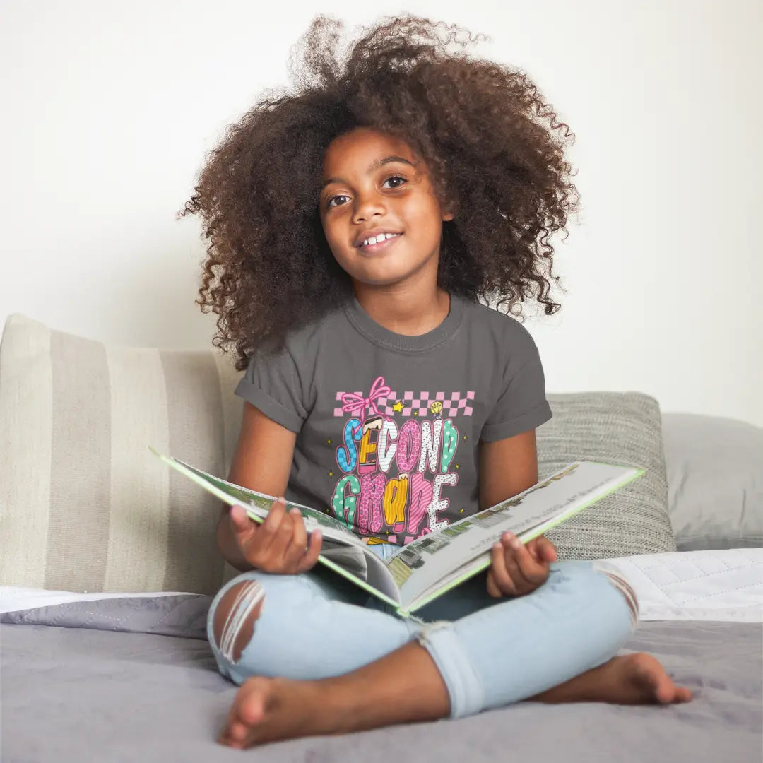Child sitting on a couch holding a book, wearing a '2nd Grade' t-shirt.