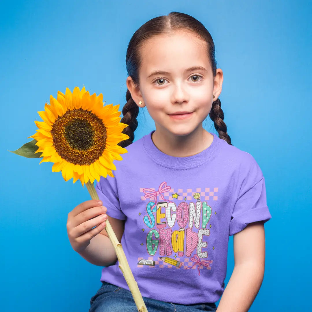 Young girl holding a sunflower against a blue background, wearing a purple 'Second Grade' shirt.