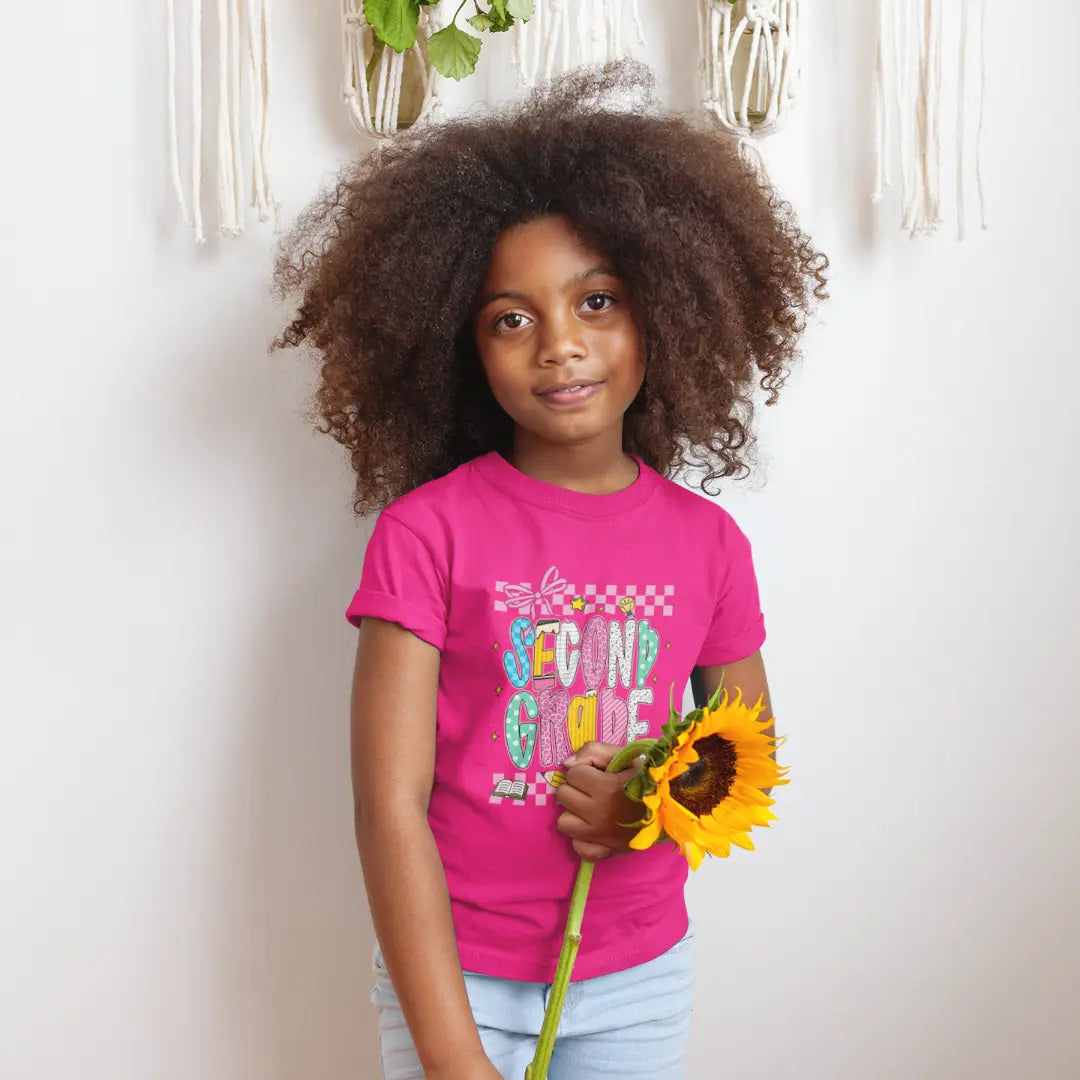 Young girl wearing a pink 'Second Grade' shirt holding a sunflower against a white wall.