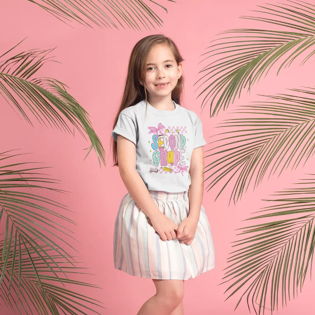 Young girl wearing a colorful second grade t-shirt with text and striped skirt on a pink background with palm leaves.